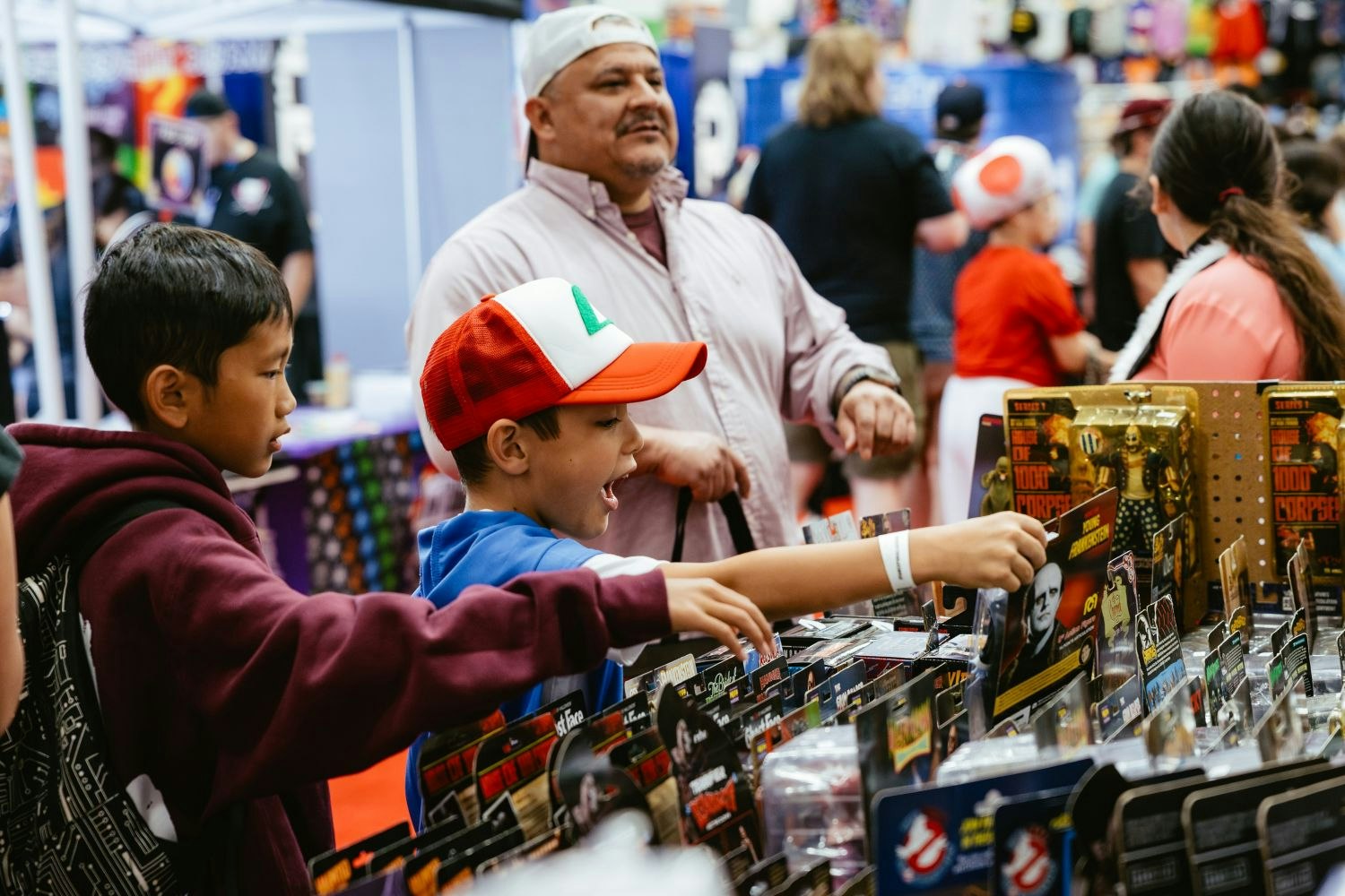 Two young fans enthusiastically point at Ghostbusters and horror-themed action figures on display, with one child wearing an Ash Ketchum cap and grinning in awe.