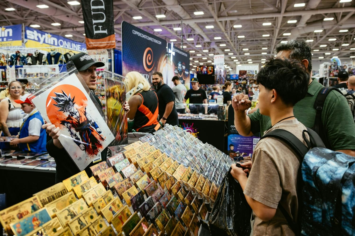 A vendor holds up anime prints for a father and son browsing a booth filled with pop culture posters and collectibles, with Dragon Ball Z artwork featured prominently.