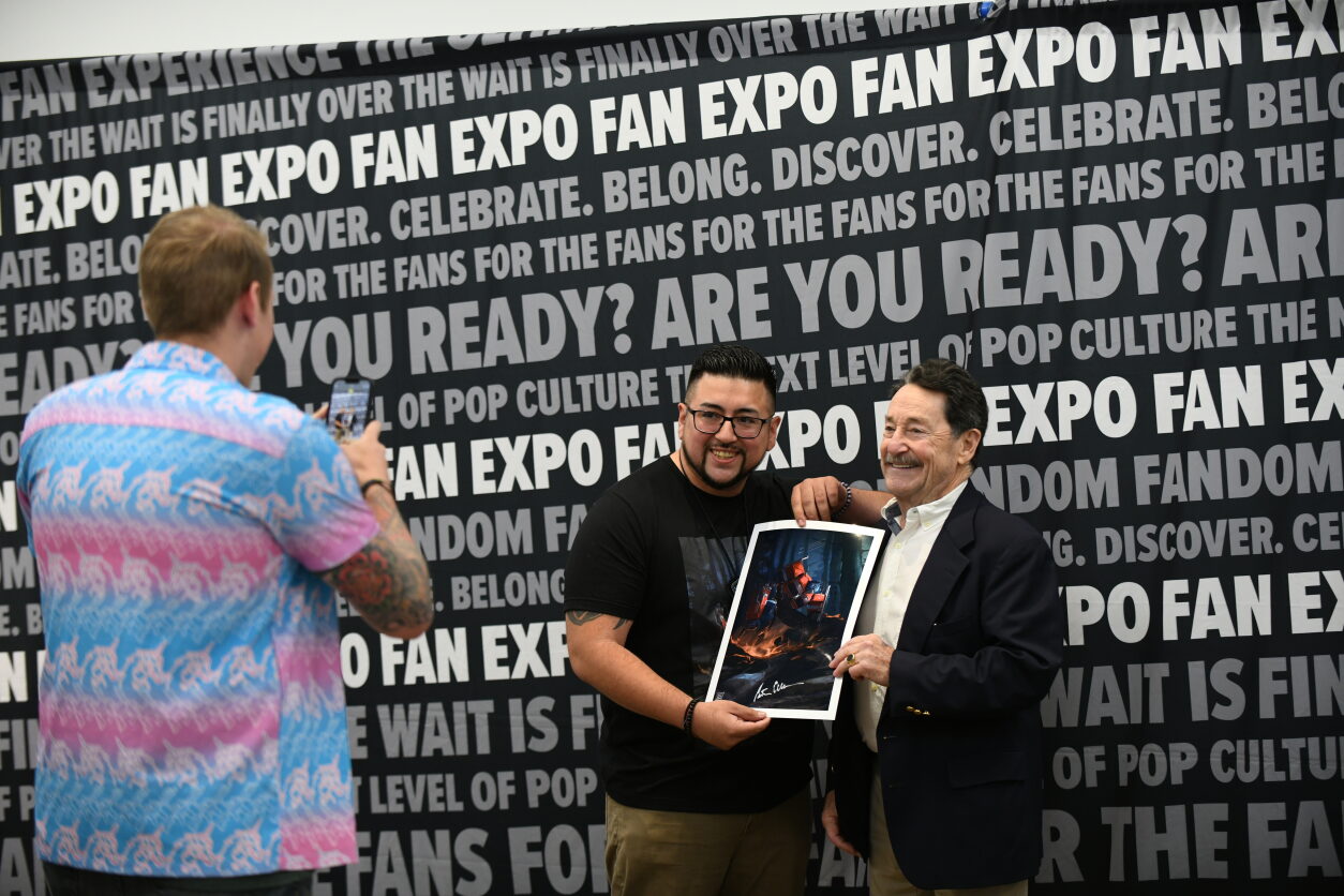 A fan gets their photo with Peter Cullen after Peter signed a hand drawn photo of optimus prime