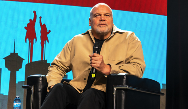 Vincent D'Onofrio is wearing a khaki coloured button up shirt with a black shirt underneath. He is sitting on stage and holding a microphone.