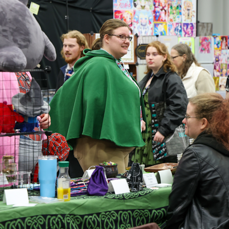 Fan dressed in a green cape admires vendor tables with stuffed toys and collectables
