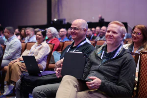 Two IT professionals attend a keynote session with laptops open, preparing to capture insights and ideas from HDI Service & Support World.