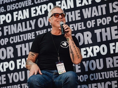 Titus Welliver, with tattoos on his arm, is speaking into a microphone, wearing sunglasses and a black t-shirt. He's seated in front of a backdrop that reads "FAN EXPO" with various other words related to pop culture. He wears a lanyard with a badge labeled "GUEST"