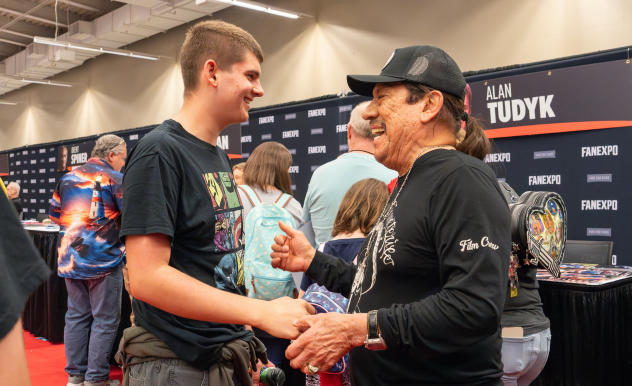 The image depicts a joyful interaction at a fan convention booth, specifically FAN EXPO. In the foreground, Danny Trejo and a fan are shaking hands and sharing a warm, engaging conversation. The younger man, smiling broadly, appears to be a fan, wearing a casual graphic t-shirt and cargo shorts. Danny Trejo is dressed in a black shirt with "Film Crew" embroidery and a cap, smiling enthusiastically. In the background, there are other attendees and promotional banners, one of which includes the name "Alan Tudyk," suggesting the event's vibrant and fan-focused atmosphere.
