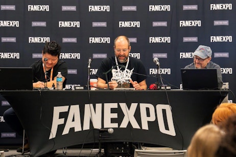 The image captures a sketch duel at FAN EXPO featuring three male panelists engaging with the audience. The man in the center, who appears focused, is handling what seems to be a detailed model or figure, while the man on the left is looking down, possibly adjusting equipment or examining materials. The panelist on the right is smiling and looking at his laptop, likely interacting with online participants or managing a presentation. The backdrop prominently displays the FAN EXPO logo, setting the professional yet fan-centered atmosphere of the panel discussion.