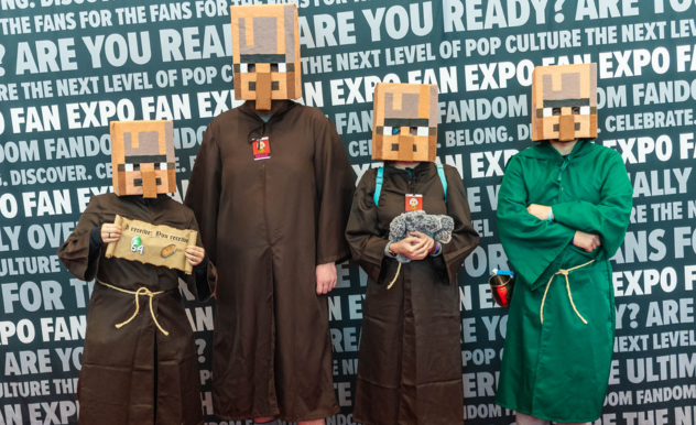 The image features four individuals standing side by side, each wearing a costume that includes a large pixelated headpiece resembling the blocky aesthetic of a popular sandbox video game. The first three are dressed in brown robes with rope belts, while the fourth person is in a green robe, holding a plush toy that looks like a grey blocky animal from the same game. They are standing in front of a backdrop that has repeated text "FAN EXPO" which suggests they are at a fan convention or event dedicated to pop culture and fandoms. The first person is holding a sign that seems to be a humorous play on the game's elements, but the exact text is not completely visible.