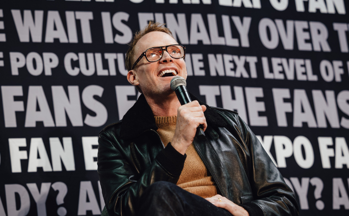 Paul Bettany with glasses and a leather jacket, holding a microphone and laughing against a backdrop with bold white text related to pop culture and fan events.