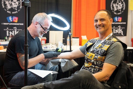 The image shows a tattoo artist at work in a convention setting. The artist, a middle-aged man with grey hair and glasses, focuses intently on tattooing the arm of a smiling, seated man wearing a graphic vest featuring movie motifs. They are surrounded by a vibrant orange backdrop with promotional banners that include logos and social media handles, indicating the professional nature of the booth. A circular light fixture provides illumination, enhancing the focus on the tattooing process, capturing a moment of creativity and skill in a public event environment.