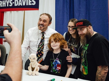 At a convention booth, four individuals are posed together for a photo. A man in a white shirt, wearing a tie with colorful characters, smiles warmly while seated behind a table displaying a figurine of a small, creature-like character. Standing to his right, a young child with curly red hair grins, flanked by a woman with glasses and long hair, and a man with a full beard and tattoos, wearing a cap labeled "FREAKSHOW." They're all sharing a cheerful moment, with a hand visible in the foreground capturing their photo on a device. Above the seated man, a sign reads "RHYS-DAVIES."