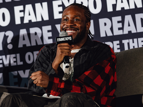 Shameik Moore smiling, sitting on a couch, talking into a microphone.