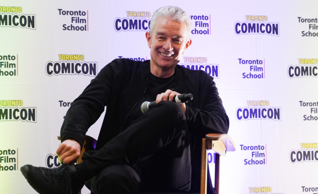 James Marsters adjusts himself in the directors chair, his lefts are crossed and he smiles, with his mic resting on his knee.