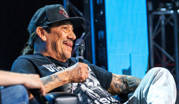 Danny is wearing a black baseball cap and a black and white graphic t-shirt. He is holding up a microphone and looking into the crowd.