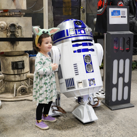 A small child in a dinosaur dress and grogu headband poses for a photo next to R2-D2