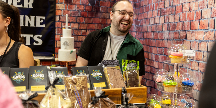 Behind a table full of various sweets like chocolate bars, cake, and cupcakes, a vendor smiles as they converse with fans.