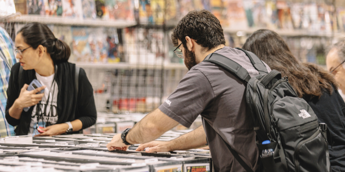 a couple of fans flip through comics in cardboard bins at a comic booth.