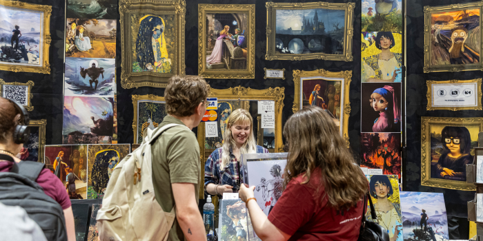 Framed by an ornate gold frame in the booth backdrop, a vendor smiles while fans shop. The booth shows famous paintings altered to have the faces of game and movie characters.