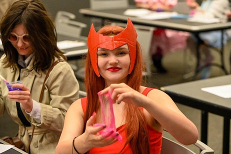 The image captures a lively scene at a convention workshop where attendees are engaged in hands-on activities. A young woman with bright red hair, wearing a vivid red dress and a matching headpiece that resembles devil's horns, playfully holds up a stretchy pink slime between her hands, smiling at the camera. Beside her, another participant, dressed in a tan trench coat and inspecting her own slime creation, appears focused and amused. The setting is informal, with participants seated at a typical conference table, reflecting the interactive and fun nature of the event.