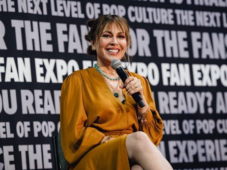 Alyssa Milano with a radiant smile is seated on stage, holding a microphone. She's wearing a mustard-colored dress with teal beaded necklaces. The backdrop features bold, repeating text emphasizing "FAN EXPO" and phrases related to pop culture. Her cheerful demeanor suggests she's engaging with an audience during a panel or interview.
