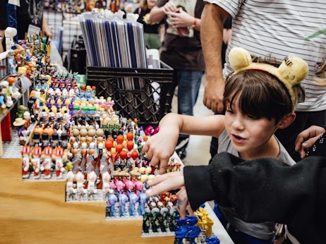 A young child with bear ear headbands excitedly reaches out to a table filled with an array of colorful miniature figures at a bustling market or convention. Surrounding patrons and stalls blur in the background, and a guardian stands closely behind the child, watching their selection. The variety of figures on display range from superheroes to animals, capturing the child's fascination.