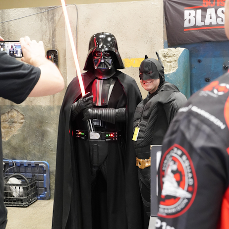 Infront of the 501st booth, a smiling Batman poses with Darth Vader, who holds his illuminated saber. A friend of Batman takes a photo on their phone.