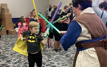 little boy wearing a batman tshirt and cape, preparing to force push their trainer, holding a foam lightsaber