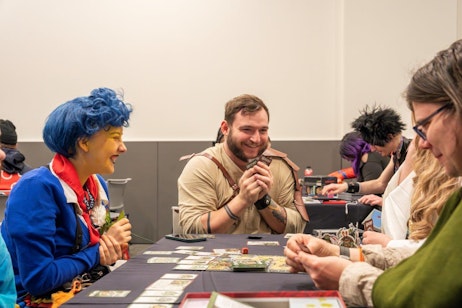 The image shows a joyful moment at a gaming table during a convention. A woman with vibrant blue hair, dressed in a blue jacket with red accents, laughs heartily, clutching a green flower. Opposite her, a man in a beige adventure vest smiles broadly, cards in hand, adding to the festive atmosphere. They are surrounded by other attendees focused on card games spread out on the table, each engrossed in the activity. The setting is casual, with a mix of colorful outfits and expressions of enjoyment, capturing the spirit of communal fun and engagement typical of fan conventions.
