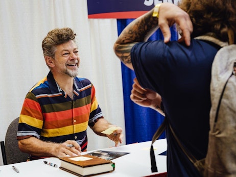 A cheerful Andy Serkis with wavy hair, wearing a vibrant multi-colored striped shirt, sits at a table with books and markers. He is engaging in a conversation and looking up towards a person with tattooed arms, whose back is partially visible in the foreground. The setting appears to be a convention or signing event with a draped white background and a glimpse of a blue banner.