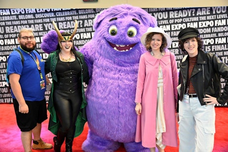 The image features a cheerful group posing together at a fan convention. From left to right: a man in a blue shirt and black shorts with a convention badge, a woman dressed in a Loki-inspired costume complete with a green cape and horns, a large, plush purple character with a big smile, a woman in a pink coat and white hat, and another woman styled in a classic black leather jacket and striped top. They are all standing on a red carpet against a backdrop with the FAN EXPO logo, visibly enjoying the lively atmosphere of the event.