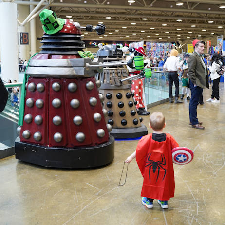 A small child follows after a pair of Daleks. The daleks are holding green beer steins and wearing green bow ties, while one has a green checkered hat. All for St. Patrick's Day. The little child has a Spider-man cape, and a Captain America shield. They are facing away from the camera.