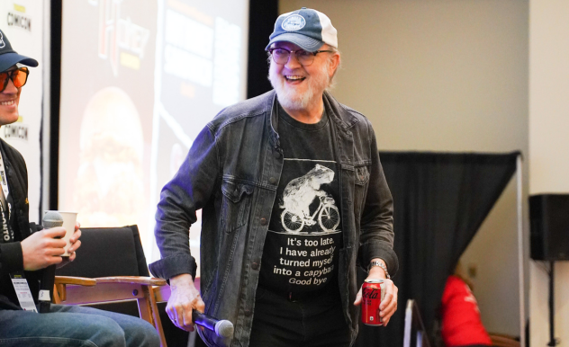 Jim Beaver stands in front of the director's chair on stage before his panel. He is holding a can of Coke Zero, and smiling at Alexander Calvert (left)