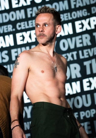 The image depicts Dominic Monaghan standing shirtless in front of a backdrop with the "FAN EXPO" logo repeated across it. He has a short, styled haircut and sports a mustache and beard. He wears green trousers and a necklace and has tattoos visible on his chest and arm. His expression is focused and slightly turned to the side, giving him a contemplative look. The overall setting suggests an event or performance at a pop culture convention.