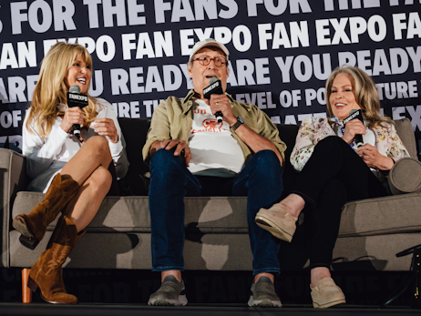 Christie Brinkley (left), Chevy Chase (middle), and Beverly D'Angelo (right) sitting on a couch together, smiling and talking together at a panel. They are all holding microphones.