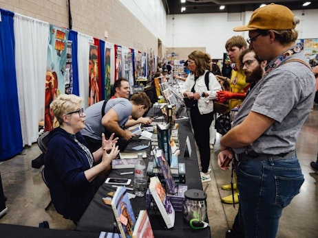 Several individuals interact at a convention booth lined with colorful banners and various items for sale. A woman with short blonde hair and glasses, wearing a dark blue top, enthusiastically talks and gestures with her hands, seated behind the booth. Two men are seen in the background engaging with their surroundings, one looking at a device and the other conversing. A group of attendees, including a man in a yellow hat and another wearing a peace-sign shirt, examine the displayed items. The setting suggests a lively and engaging comic or pop culture convention.