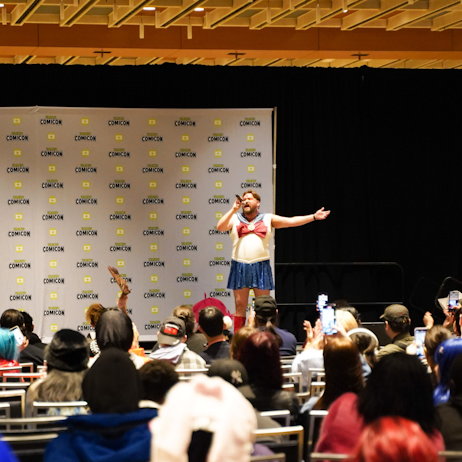 Bear Sailor Moon (a male drag performer with short brown hair and a beard, dressed as sailor moon) sings with and outstretched arm to a large crowd