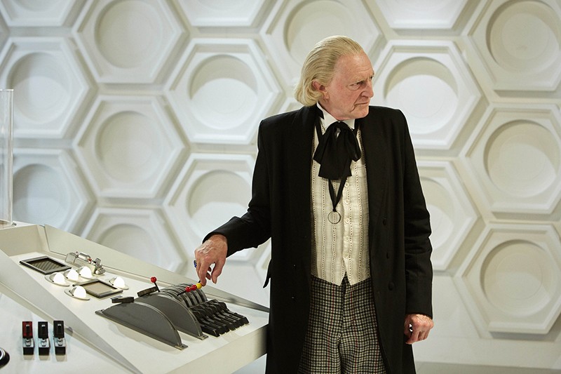 The Doctor stands next to the Motherboard of the TARDIS, reaching for the one yellow lever along a row of red levers. He stares sternly off screen.