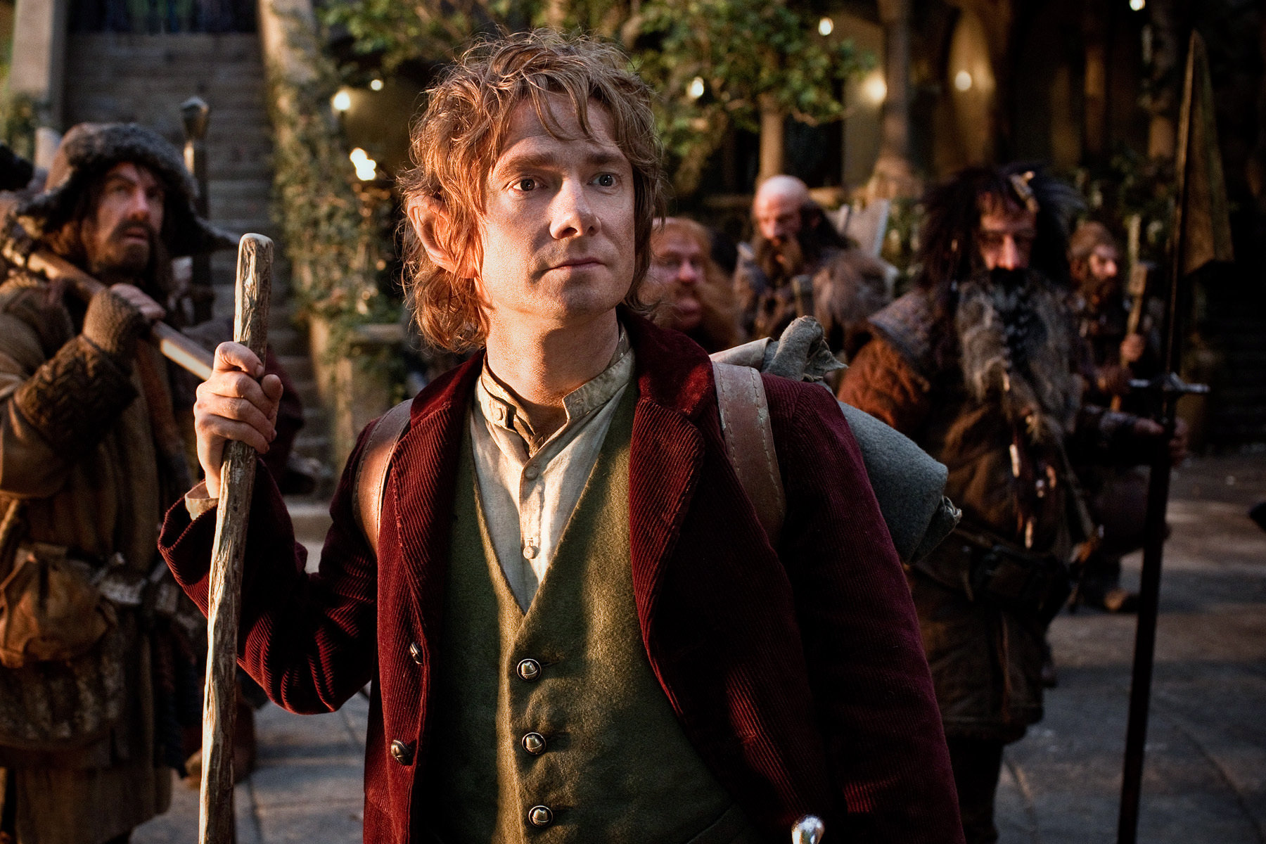 Bilbo Baggins stands there looking off into the distance. He is wearing a white button up shirt, a green vest and a burgundy corduroy jacket and holds a wooden walking stick. Other hobbits stand behind him in the background.