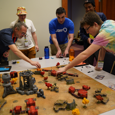 5 people stand table side. on the table is miniatures for Battlefront. 2 people are moving miniatures, one is picking up play cards as the other two watch