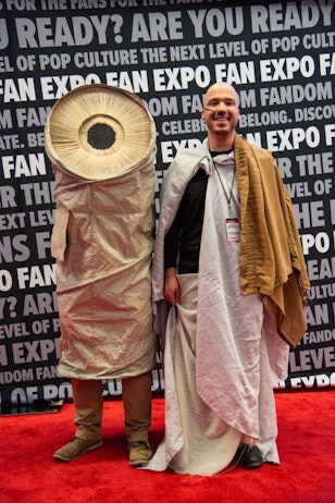 The image shows two people at a FAN EXPO event, posing against a backdrop with event logos. On the left, one person is dressed in a creative costume that resembles a worm from “DUNE”, featuring a large, detailed iris and pupil on a round, beige-colored body. The person on the right is smiling broadly, bald, and dressed in a Jedi-like costume with a long, white robe. They are standing on a red carpet, contributing to the festive atmosphere of the event.