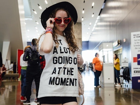 A young woman with curly hair stands confidently in an indoor venue, adjusting heart-shaped sunglasses. She wears a black hat and a white T-shirt with the bold text "A LOT GOING ON AT THE MOMENT". In the background, attendees can be seen milling about, with informational booths and staff present. The atmosphere is lively, suggesting a convention or large gathering.