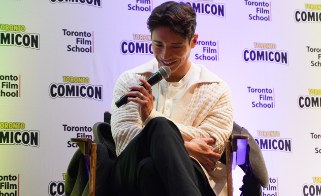 Manny Jacinto is seated in a director's chair with his legs crossed. He leans forward, smiling into his microphone.