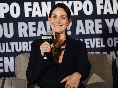 Carrie-Anne Moss sitting on a chair on a stage, smiling and holding a microphone.