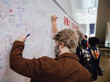 People are gathered at an indoor event, signing and leaving messages on a large white wall filled with colorful handwriting and doodles. A man in the foreground, wearing a brown jacket, writes on the wall with a blue pen. The crowd behind him includes a person in a detailed jacket with badges and an embroidered phoenix. The wall, acting as a communal canvas, captures the energy and contributions of the attendees.