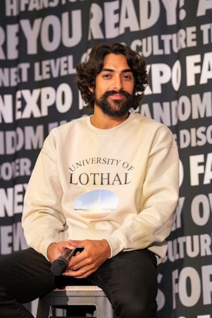 The image captures Eman Esfandi seated on a stool at a panel event, holding a microphone. He has shoulder-length curly hair, a full beard, and a thoughtful expression. He is dressed casually in a cream-colored sweatshirt with "University of Lothal" printed on it, and black pants. The backdrop features a repeated text pattern reading "FAN EXPO," emphasizing the event's focus on pop culture. His relaxed posture and attentive gaze suggest he is engaged in a discussion or preparing to speak.