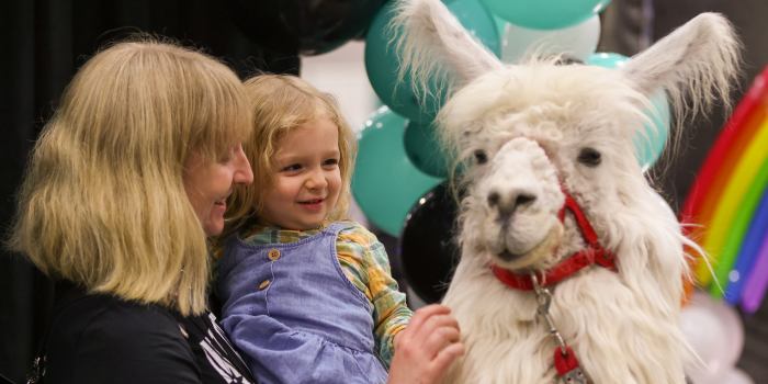 A MOTHER AND DAUGHTER SMILE, SHARING A SWEET MOMENT WITH LOCAL LLAMA, CAESAR, IN FRONT OF THE COMMUNITY ZONE BALLOON ARCH