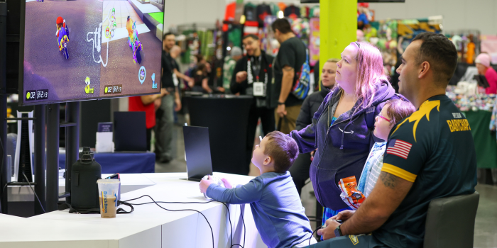 MARIO KART IS A FAMILY AFFAIR AT THE US NAVY BOOTH. A YOUNG CHILD PLAYS MARIO KART AGAINST A MEMBERS OF THE GOATS AND GLORY US NAVY ESPORTS TEAM WHILE HIS MOTHER AND SIBLING SPECTATE.