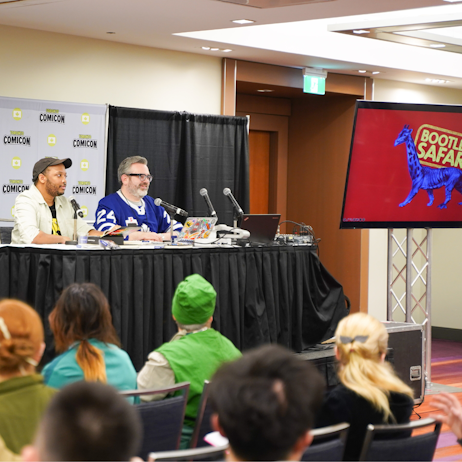 Aaron Reynolds, with glasses, greying hair and facial hair, sits right, in a Toronto Maple Leafs jersey, left at the table is Paris Alleyne, in a black tshirt, beige overshirt, and grey hat. The screen beside them reads Bootleg Safari, and has an image of an animal with lion body and giraffe head.