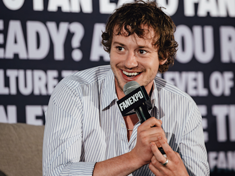 Joseph Quinn smiling and holding a microphone.