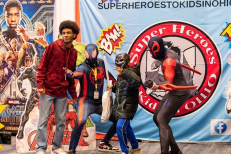 The image features a dynamic group posing at a comic convention. On the left, a young man stands in casual attire, with a large afro and a red hoodie, alongside three children dressed in superhero costumes. The kids embody characters from popular comics: Spider-Man with a modern twist, a futuristic knight, and a red ninja character. They're positioned in front of a colorful backdrop promoting superhero-themed initiatives for kids, complete with comic book-style graphics and logos. The group's playful stances and costumes add a lively, festive atmosphere.