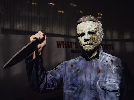 A person dressed as a horror movie character, wearing a decaying mask and tattered blue jumpsuit, menacingly holds up a knife. The background features a dimly lit wall with a sign that reads "WHAT'S YOUR FAVORITE DALLAS." The ominous lighting casts shadows, enhancing the eerie atmosphere of the scene.