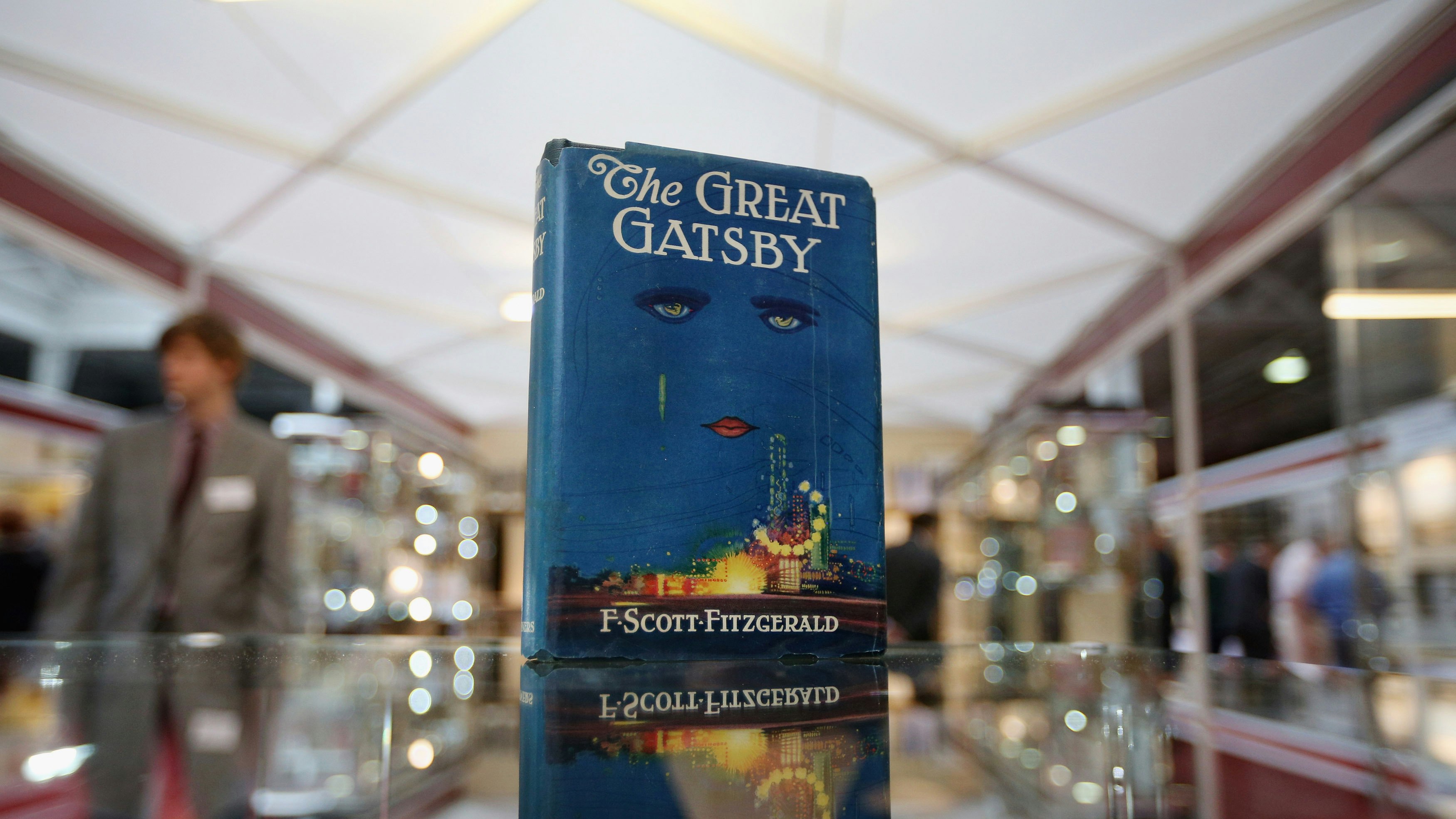 A copy of The Great Gatsby sits in a room full of display cases. The cover is a faded dark blue, and features the eyes and lips of a woman, with a lighthouse flashing along the bottom.